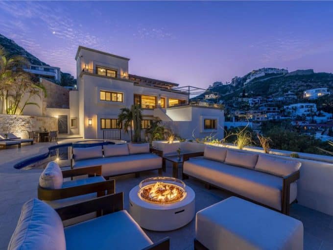 Luxury Pedregal Estate Illuminated At Dusk With Modern Outdoor Seating And A Fire Pit, Overlooking Hillside Homes And Scenic Mountain Views, Perfect For Relaxing And Entertaining.