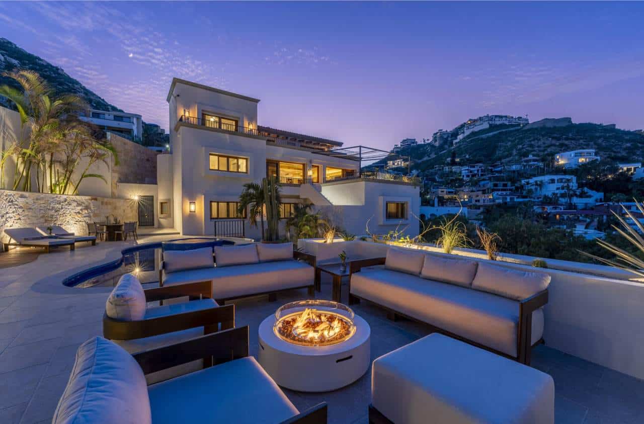 Luxury Pedregal Estate Illuminated At Dusk With Modern Outdoor Seating And A Fire Pit, Overlooking Hillside Homes And Scenic Mountain Views, Perfect For Relaxing And Entertaining.