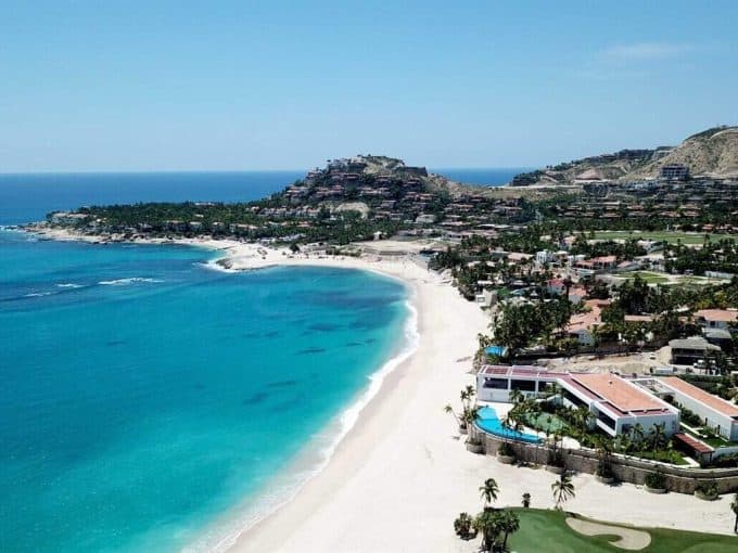 Aerial View Of Pedregal Beachfront Estate With White Sandy Beach, Turquoise Waters, Luxury Homes, And Lush Greenery In A Scenic Coastal Setting.