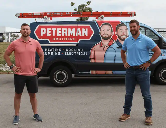 Two men stand in front of a Peterman Brothers service van with a ladder on top and a large company logo featuring illustrated portraits of two men.