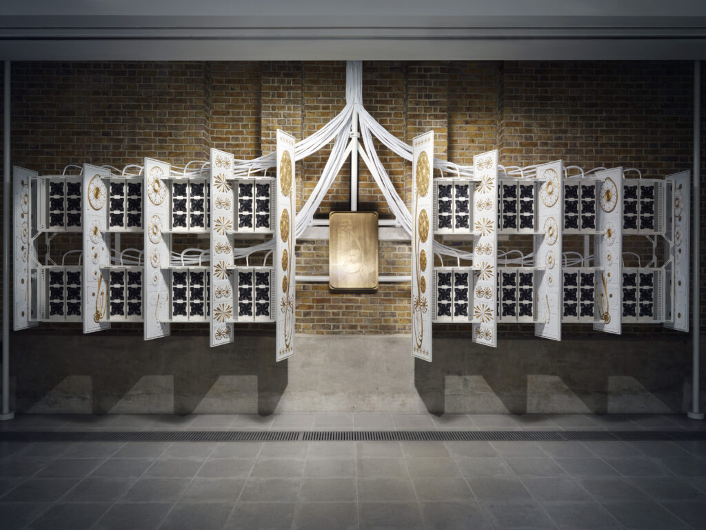 A contemporary art piece featuring a symmetrical structure with white panels embellished with gold patterns, suspended curves, and black grid elements, set against a brick wall.