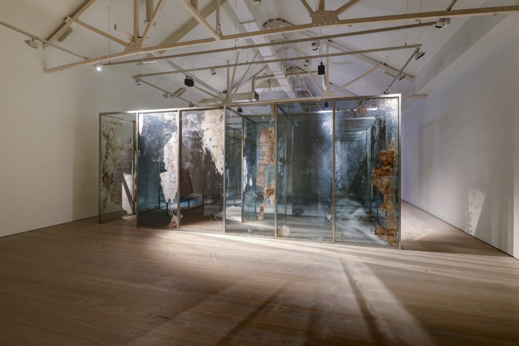 Intriguing art installation featuring weathered, partially mirrored glass panels with rust and peeling effects, displayed in a spacious, well-lit gallery.