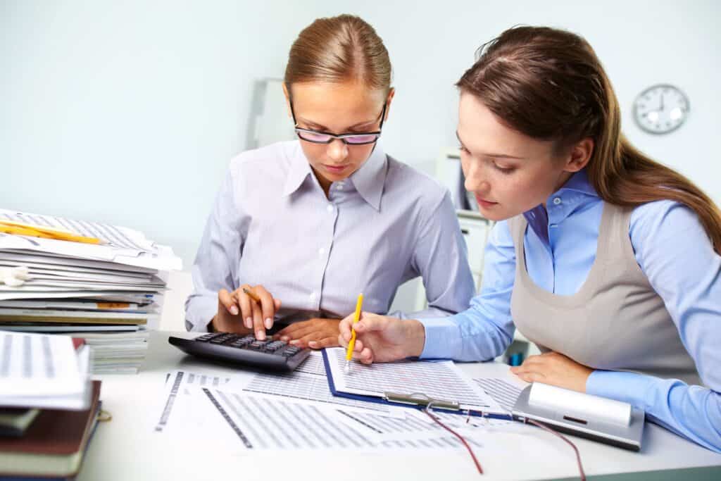 Two women working on financial documents at a desk with calculator and papers.