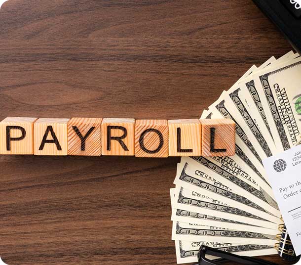 Payroll concept with wooden blocks spelling "PAYROLL" and US dollar bills on a wooden desk.