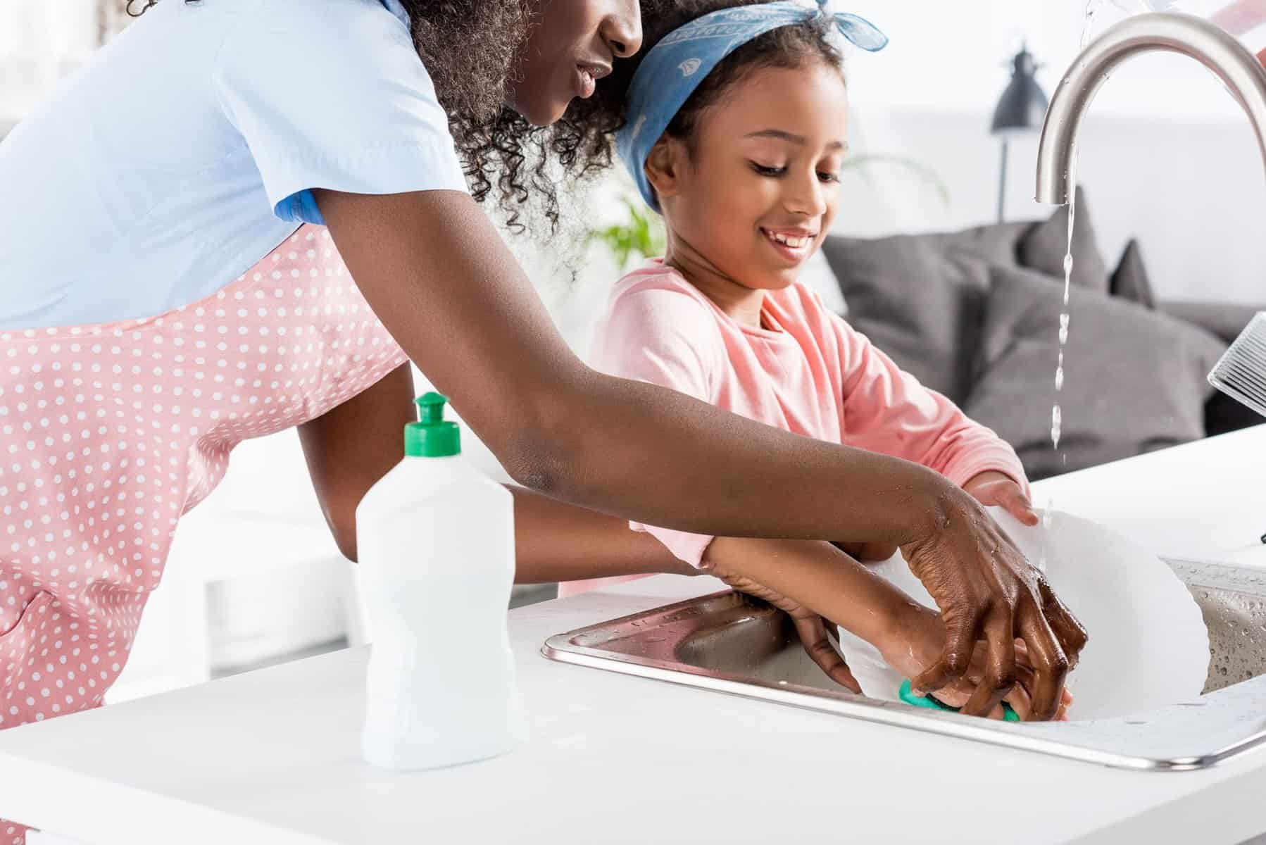 An adult and child wash dishes together at a kitchen sink, with soap and water supplied by the best home water heater.