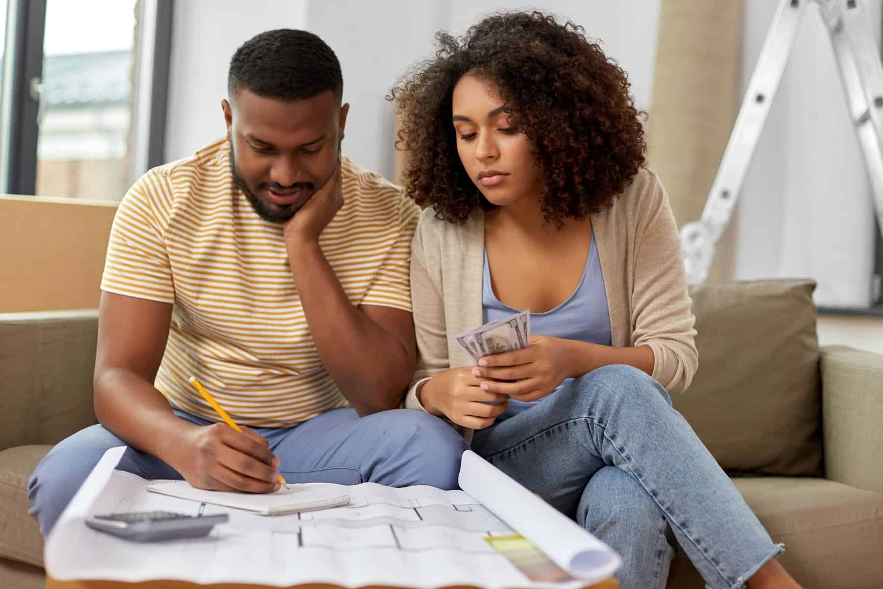 A man and woman are seated on a couch, immersed in reviewing a blueprint. The man, pencil in hand, sketches ideas while the woman holds money, contemplating the cost of hiring a professional for their HVAC system upgrade. A calculator sits nearby.