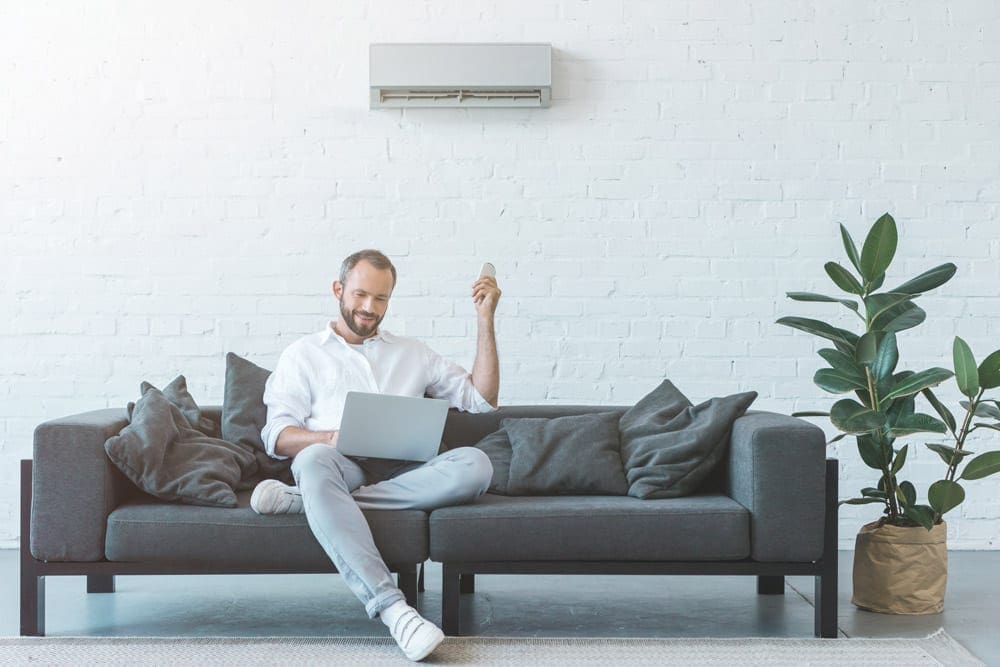 A man sits on a gray sofa using a laptop, holding a smartphone, with a potted plant nearby. Behind him, a white air conditioning unit keeps things cool for those hot summer days without skyrocketing air conditioning costs.