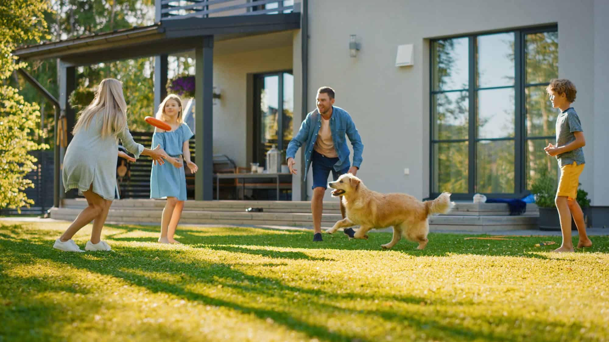 The family enjoys a game of frisbee on the grassy lawn beside their modern house, where an efficient HVAC system keeps seasonal allergies at bay. Under a clear sky, two adults, two children, and a golden retriever revel in carefree play.