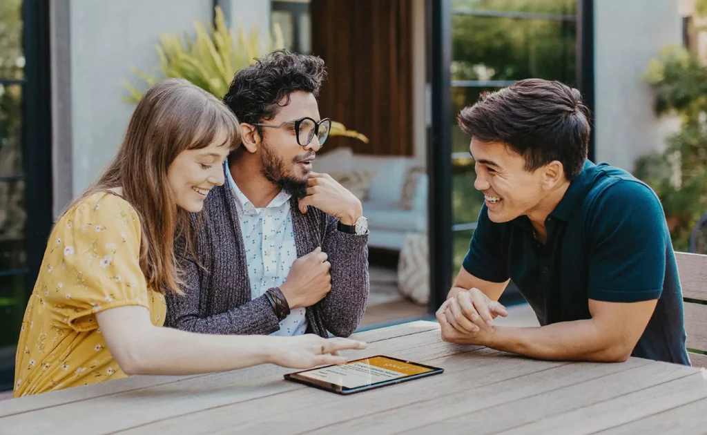 Three people sitting at an outdoor table, smiling and discussing something on a tablet.