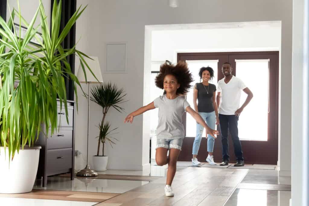 A child joyfully runs indoors towards the camera, with two adults smiling in the background. Large green plants decorate the room, kept cool by the best home air conditioning system.
