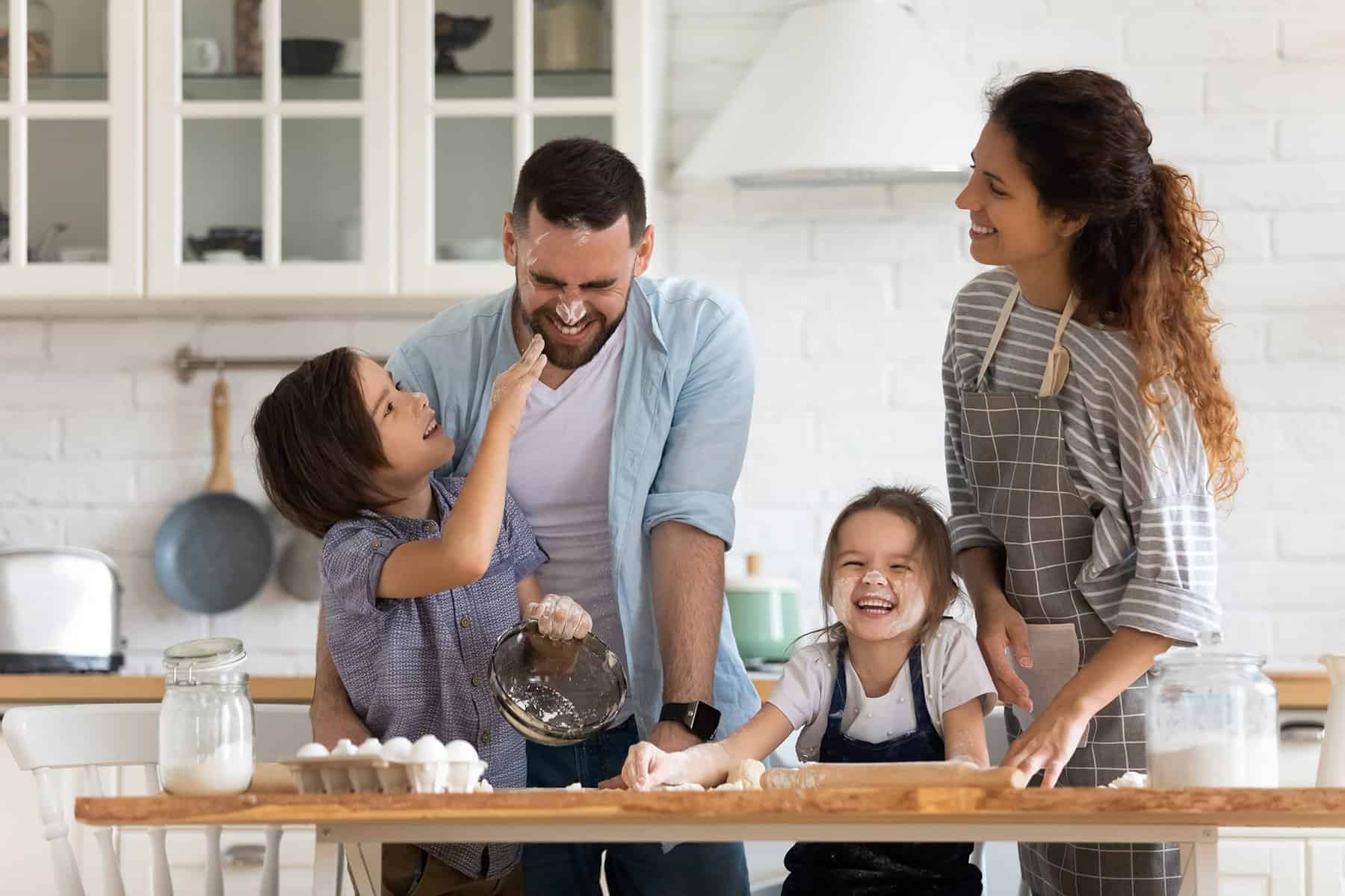 A family of four is baking in a kitchen. As one child playfully smears flour on the man's nose, the woman notices a shiny row of copper pipes along the wall, reminding her that they've put off checking for a pinhole leak amidst their laughter and dough-filled fun.