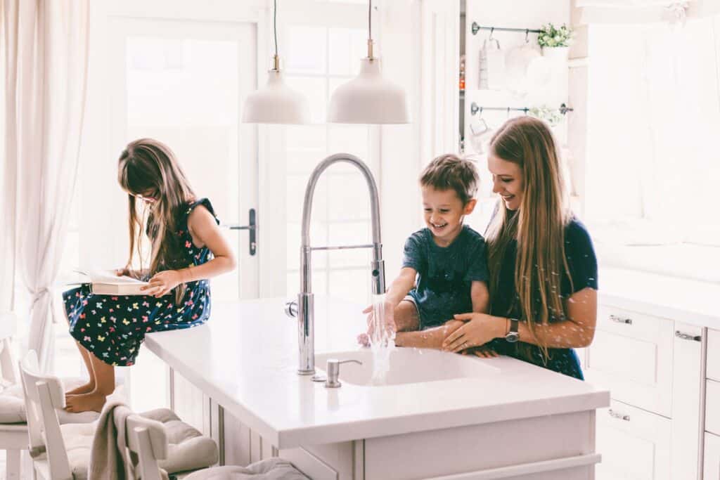 A woman and a boy play with water at a kitchen sink, enjoying the moment while subtly showcasing their functional home plumbing. Nearby, a girl sits on the counter reading, perhaps exploring maintenance tips or consulting a trusted plumbing checklist.