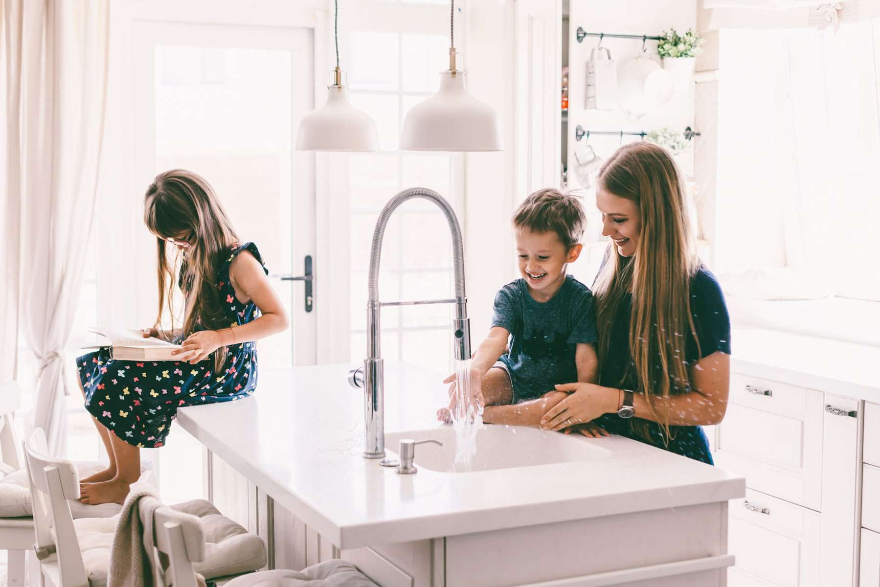 A woman and a boy play with water at a kitchen sink, enjoying the moment while subtly showcasing their functional home plumbing. Nearby, a girl sits on the counter reading, perhaps exploring maintenance tips or consulting a trusted plumbing checklist.