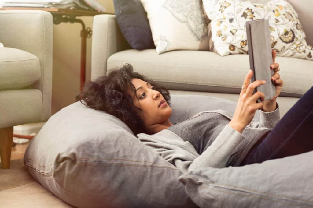 A person is lounging on a bean bag, using a tablet in a cozy room with gray couches and decorative pillows, perhaps researching an AC system upgrade to enhance comfort for those laid-back moments.