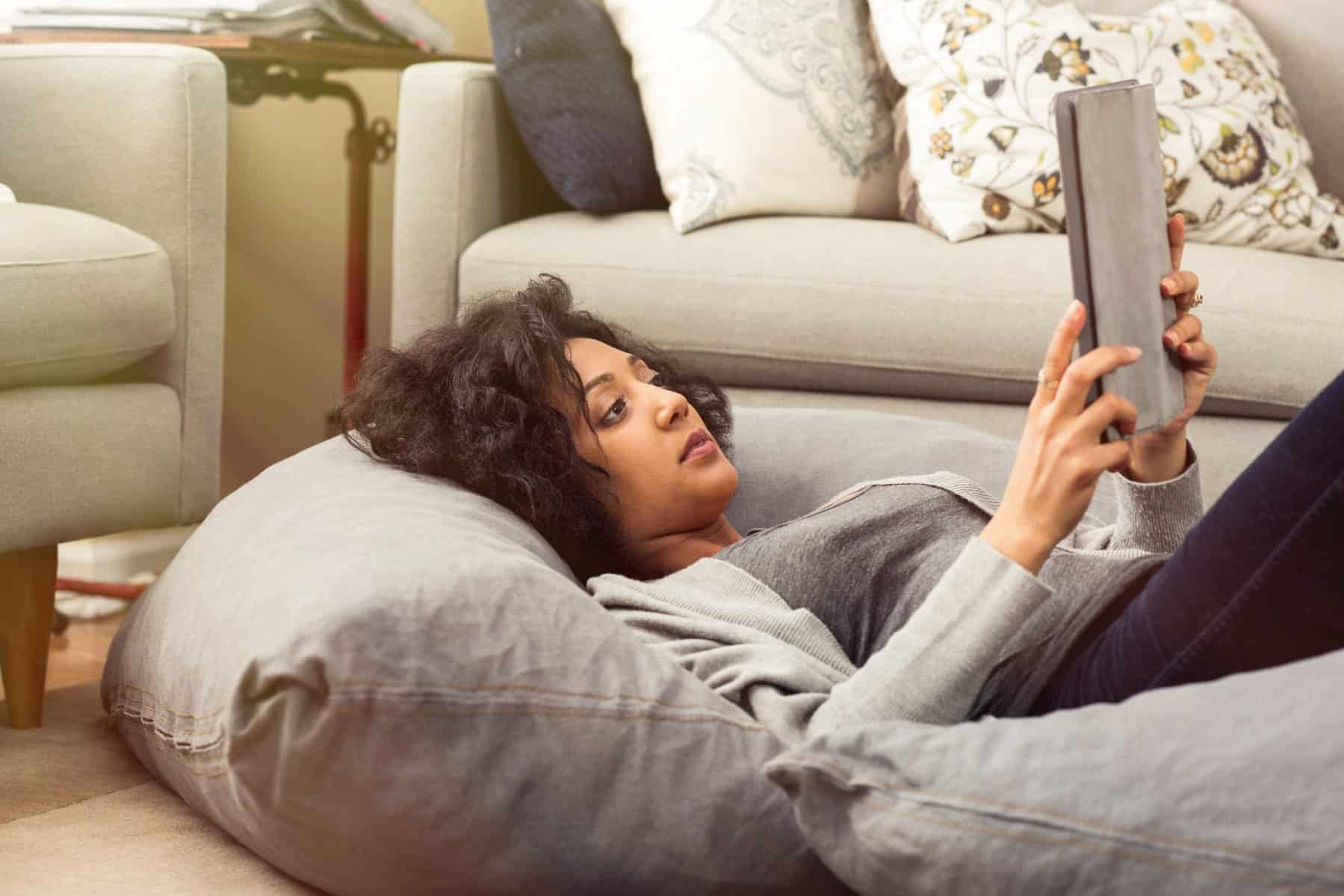 A person is lounging on a bean bag, using a tablet in a cozy room with gray couches and decorative pillows, perhaps researching an AC system upgrade to enhance comfort for those laid-back moments.