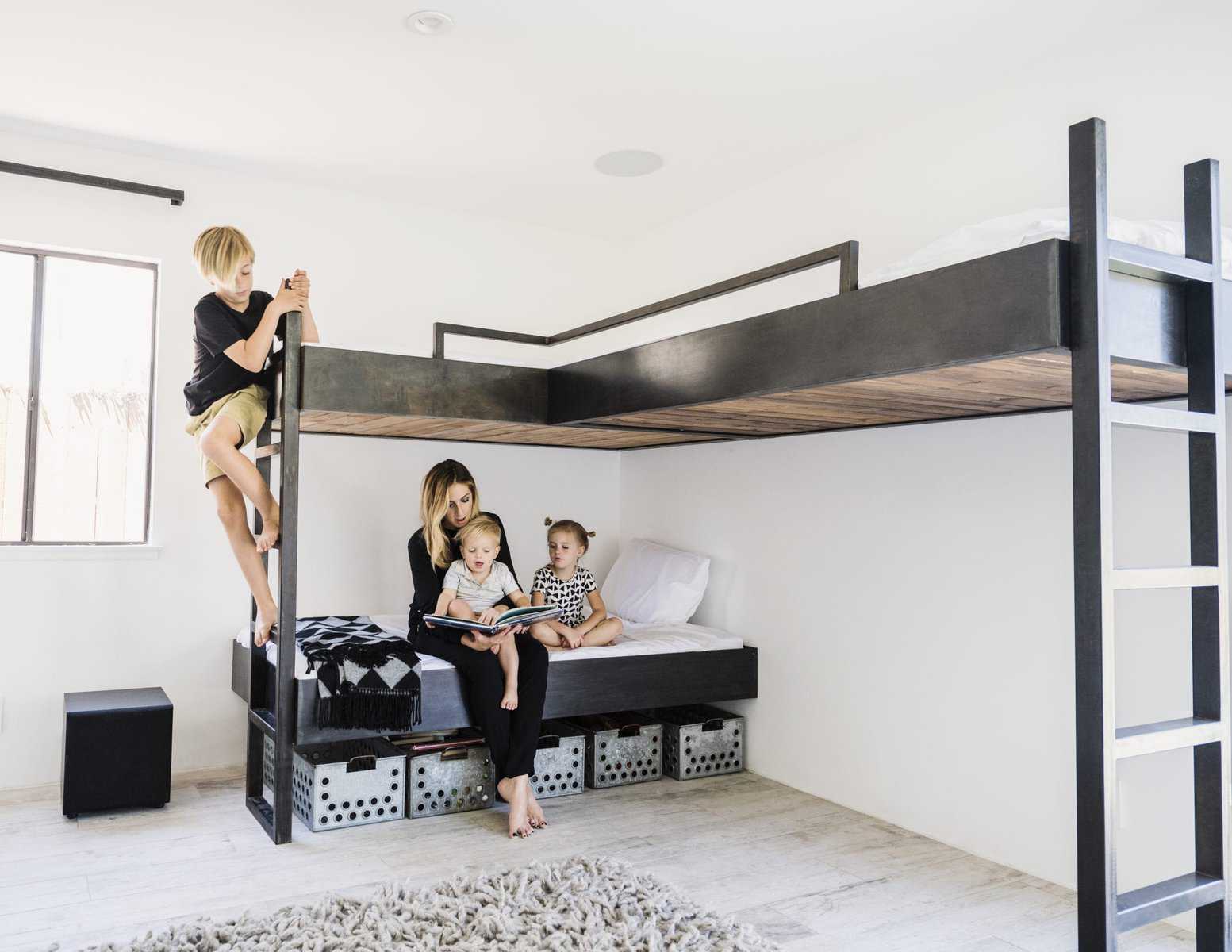 A woman sits with two young children on a lower bed under a lofted bed. A child climbs a ladder to the top bunk in their bright, minimalist room, designed with home ventilation in mind for healthier indoor air.