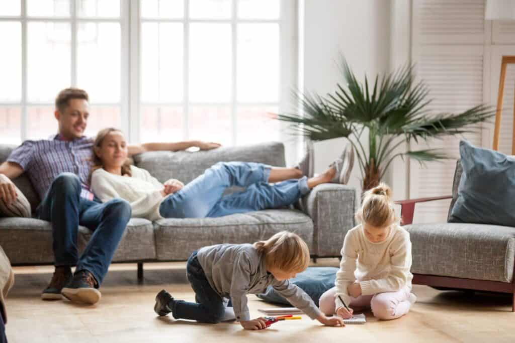 Two children draw on the floor with crayons, while a man and woman relax on a gray sofa in a bright room featuring large windows and a potted plant. Their peaceful relationship is enhanced by home humidifiers, promoting health and comfort for the whole family.