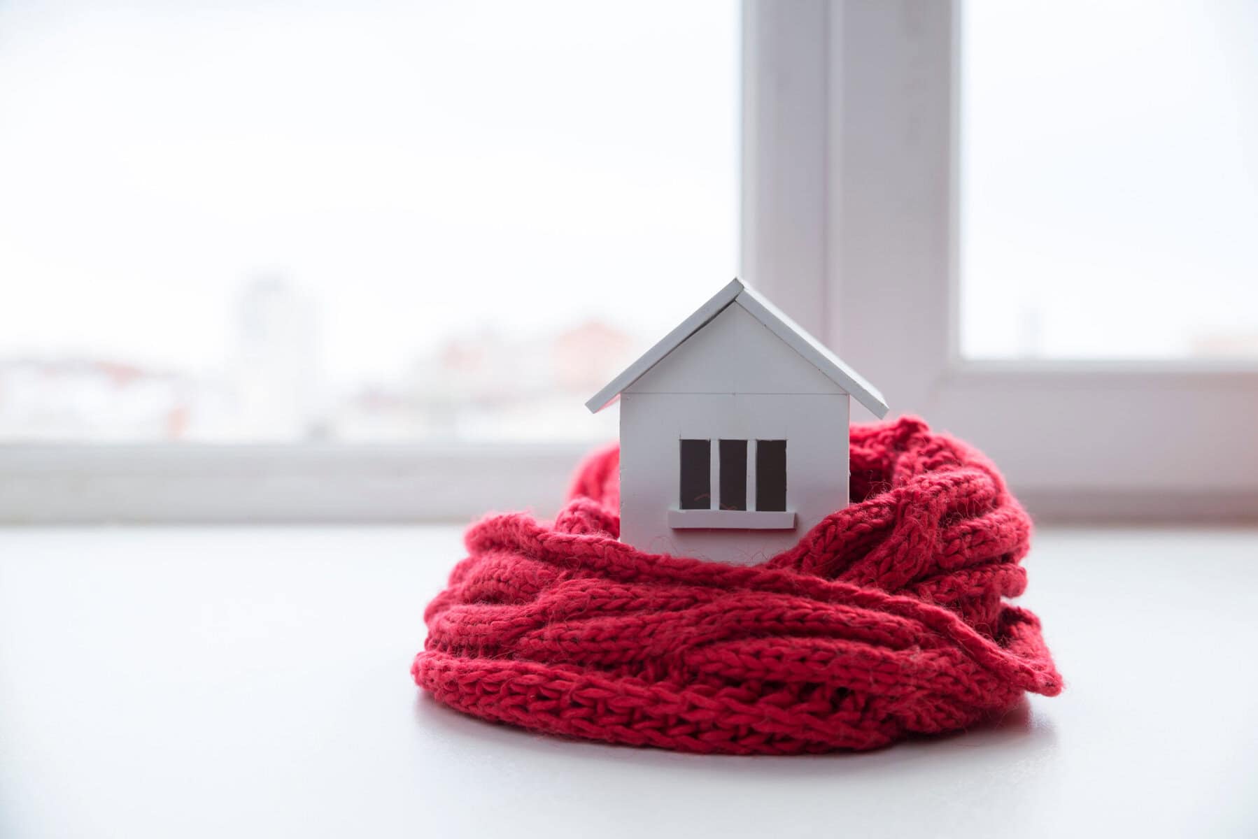 A small white house model, symbolizing efficient winter energy-saving tips, sits wrapped in a cozy red knitted scarf on a windowsill, basking in the gentle light streaming through the window.