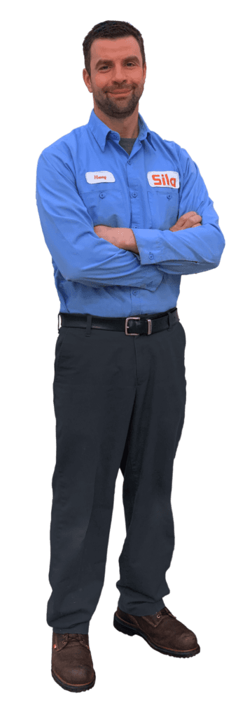 A person in a blue work shirt with a "Sila" logo and name tag stands with arms crossed, wearing dark pants and brown shoes.