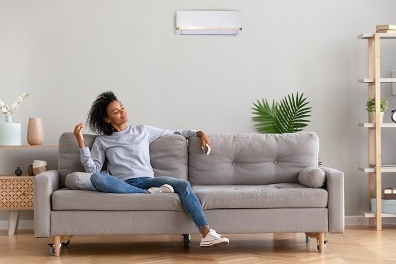 Woman sitting relaxed on a gray sofa in a modern living room, holding a remote control, with an air conditioner mounted on the wall above her—enjoying comfort thanks to reliable HVAC services.