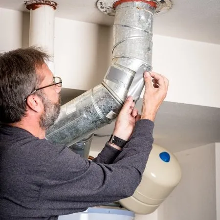 A man repairs a metal air duct by sealing a joint with tape in a utility room.