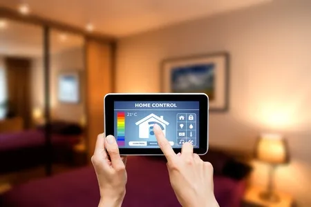 A person uses a tablet with a home control interface to adjust settings in a modern, softly lit bedroom.