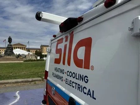 The side of a Sila service van displaying the company’s logo and services for heating, cooling, plumbing, and electrical, parked outdoors near a historic building.