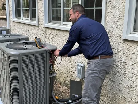 A person in a blue shirt is servicing an outdoor air conditioning unit with tools and testing equipment beside a building.