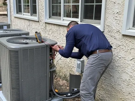 A technician is inspecting and repairing an outdoor air conditioning unit next to a house, using a digital multimeter to check the system.