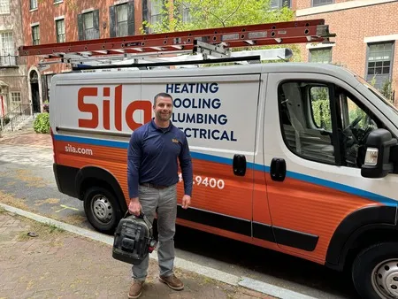 A man in a work uniform stands next to a Sila service van labeled “Heating, Cooling, Plumbing, Electrical,” holding a tool bag on a city street.