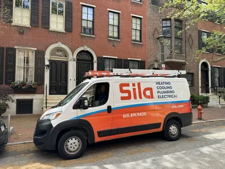 A Sila service van labeled "Heating, Cooling, Plumbing, Electrical" is parked on a residential street in front of brick townhouses.