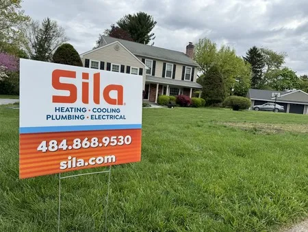 A Sila service sign advertising heating, cooling, plumbing, and electrical is displayed on the lawn of a suburban house.