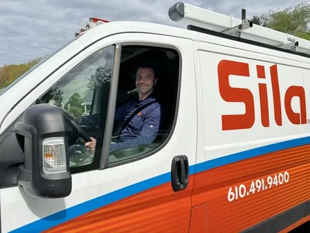 A man sits in the driver's seat of a Sila service van with a ladder on top and company contact information visible on the side.