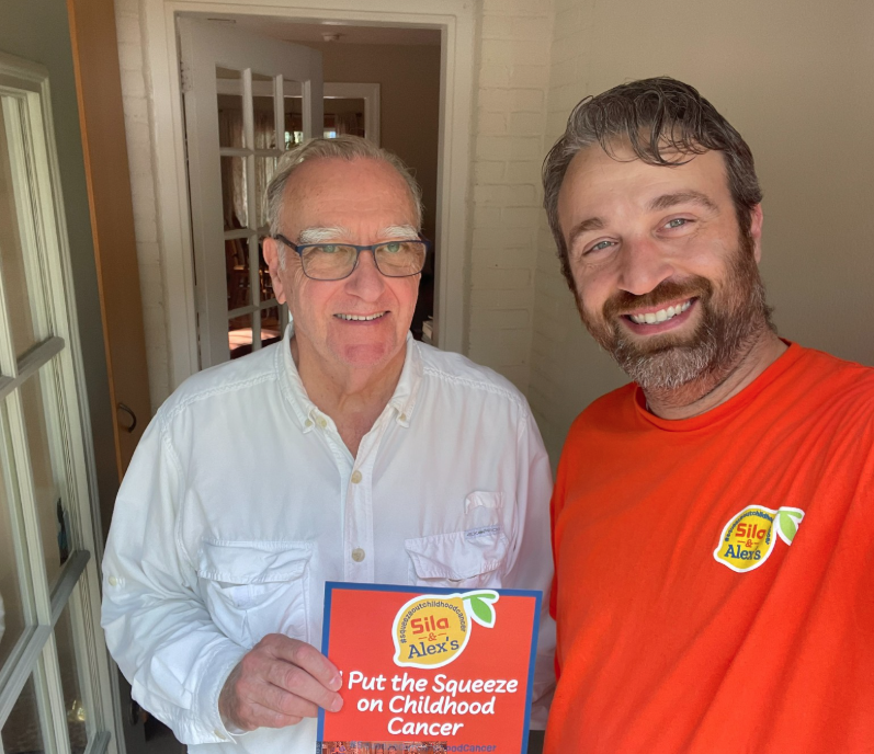 Two men stand indoors. One holds a sign reading “Put the Squeeze on Childhood Cancer.” Both wear Alex’s Lemonade stickers in support of the Alex’s Lemonade Stand Foundation.