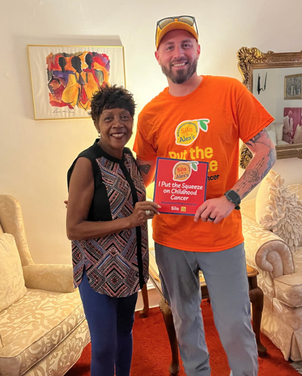 Two people stand indoors, smiling at the camera. The man holds a sign about childhood cancer awareness and Alex’s Lemonade, highlighting their commitment to the cause. The room has cream-colored furniture and artwork on the walls.