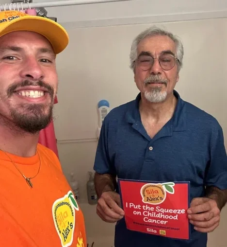 Two men stand indoors; one, wearing an orange shirt and hat, smiles warmly, while the other holds a sign reading, “I Put the Squeeze on Childhood Cancer,” showing their support for Alex’s Lemonade and its mission to fundraise for childhood cancer research.