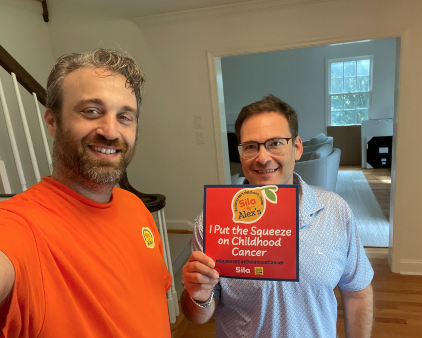 Two men stand indoors; one holds a sign reading “I put the squeeze on childhood cancer.” Both are smiling, and the background shows a room with boxes and a window, hinting at their involvement in Alex’s Lemonade efforts to fight childhood cancer.