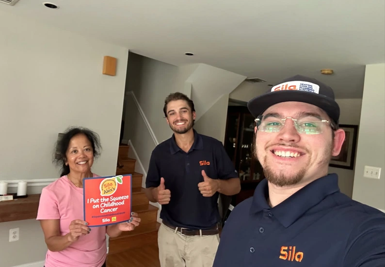 Three people stand indoors smiling; one holds a sign reading “I put the squeeze on childhood cancer” in support of Alex’s Lemonade. Two wear navy shirts and hats with “Sila” logos.