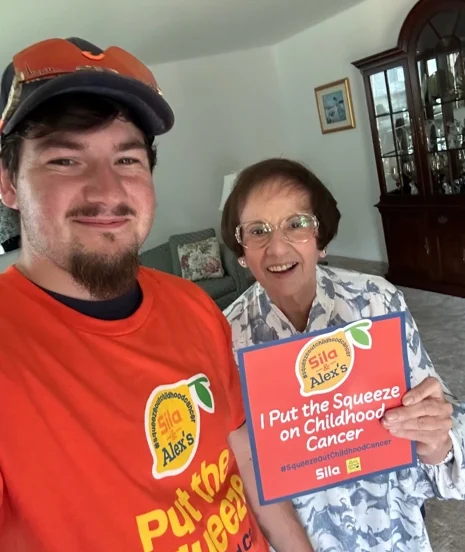 Two people smile in a living room; one wears an orange shirt and cap, while the other holds a sign reading “I Put the Squeeze on Childhood Cancer,” showing their support for Alex’s Lemonade.