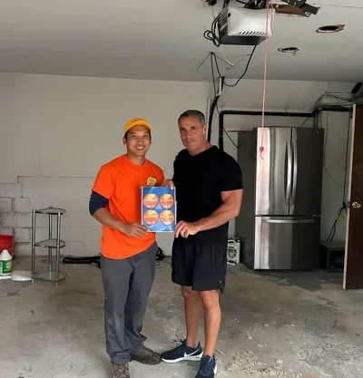 Two men stand in a garage, one wearing an orange shirt and cap, holding a colorful Alex’s Lemonade booklet—a symbol of the fight against childhood cancer. The other man wears black athletic clothes. A refrigerator and exposed ceiling are visible in the background.