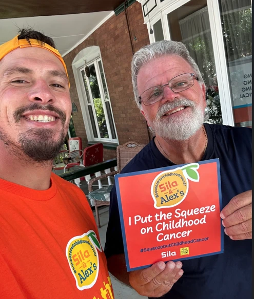 Two men smile at the camera; one holds a sign reading “I Put the Squeeze on Childhood Cancer.” Both wear orange shirts proudly displaying the Alex’s Lemonade logo.
