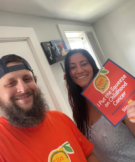 Two adults smile indoors; one holds a sign reading “I Put the Squeeze on Childhood Cancer” for an Alex’s Lemonade awareness campaign.