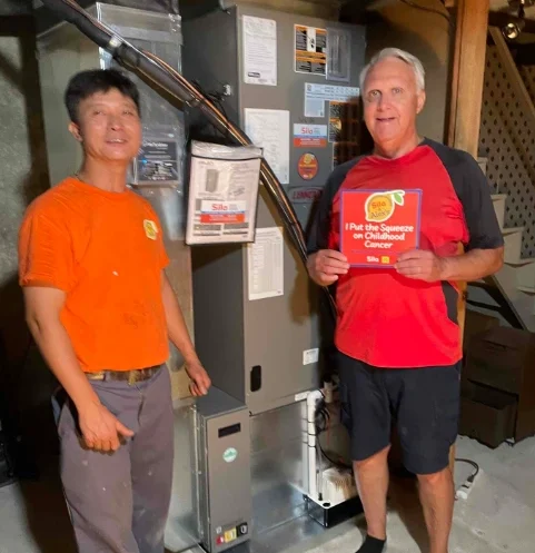 Two men stand in a basement next to an HVAC unit; one man holds a sign that says, “I Put the Squeeze on Childhood Cancer!”—showing their support for Alex’s Lemonade in the fight against pediatric cancer.