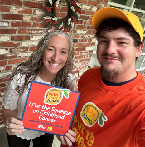 Two people smile at the camera; one holds a sign supporting childhood cancer awareness and Alex’s Lemonade charity. Both wear bright shirts and a hat with a porch and brick wall in the background.