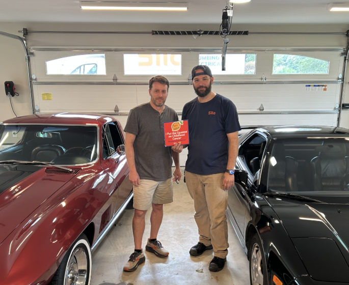 Two men stand in a garage between a red classic car and a black sports car, holding a red book and facing the camera, united in support of Alex’s Lemonade—a charity dedicated to fighting childhood cancer.