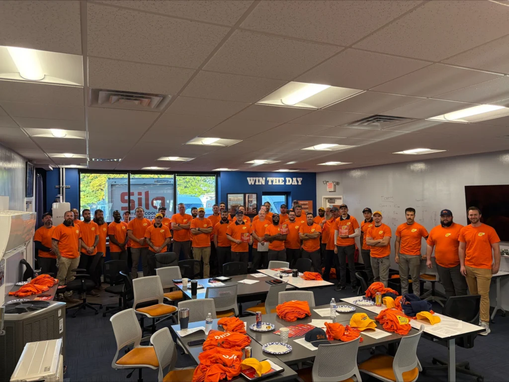 A group of people in matching orange shirts stand in a conference room behind tables with snacks, shirts, and hats, participating in an Alex’s Lemonade fundraiser.