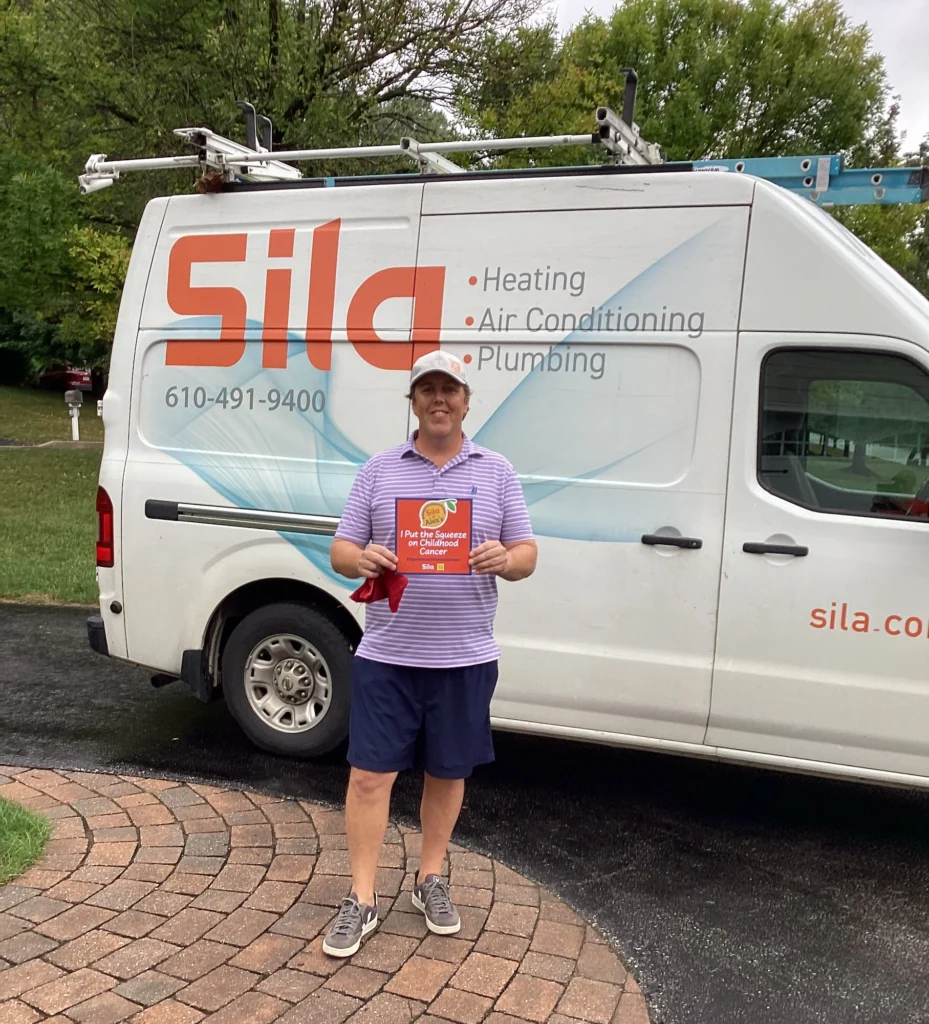 Person in a striped shirt and shorts stands on a brick walkway holding an Alex’s Lemonade sign, in front of a white Sila service van parked on a driveway.