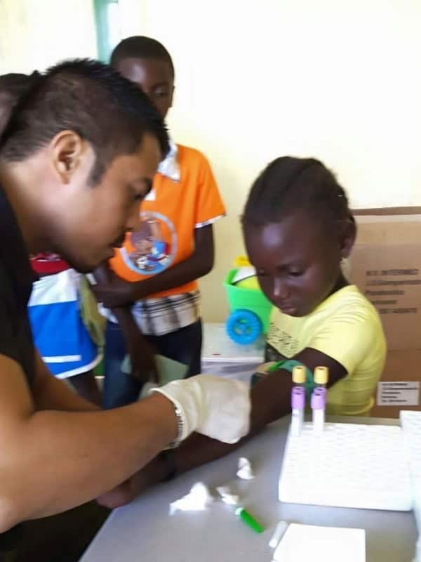 Doctor wearing a medical glove helps a young girl during a community health visit while other children look on.