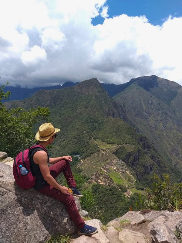 Hiker sitting on a rocky overlook above Machu Picchu and the surrounding green Andes mountains.