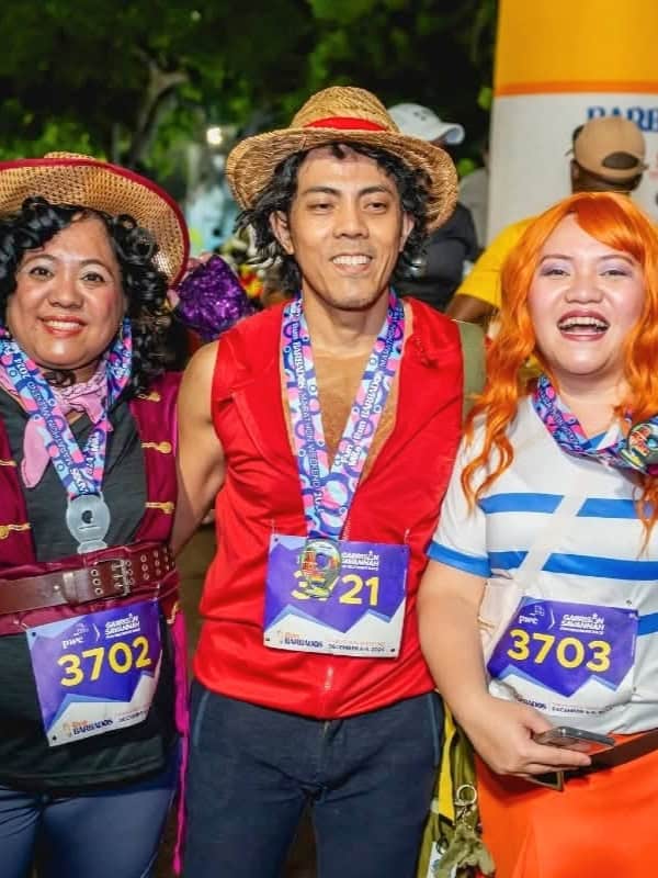 Three people dressed as One Piece characters pose together at a race event wearing straw hats and colorful race bibs.